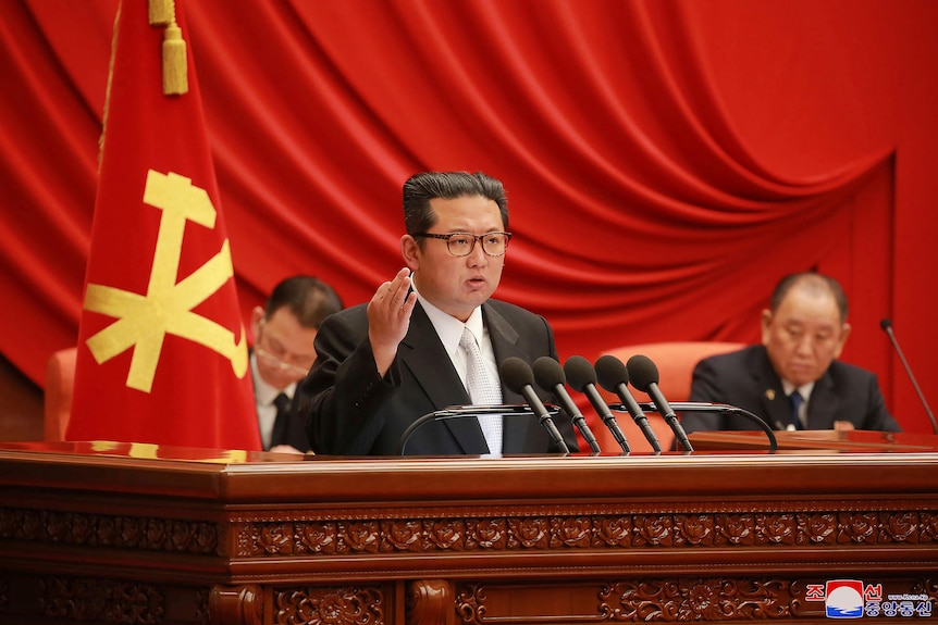 Kim Jong Un raises his hand while speaking at a large wooden desk.