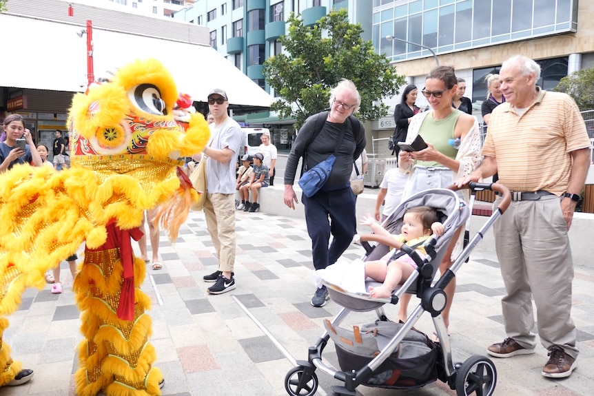 A yellow lion costume approaches standing audience members and a baby in a pram