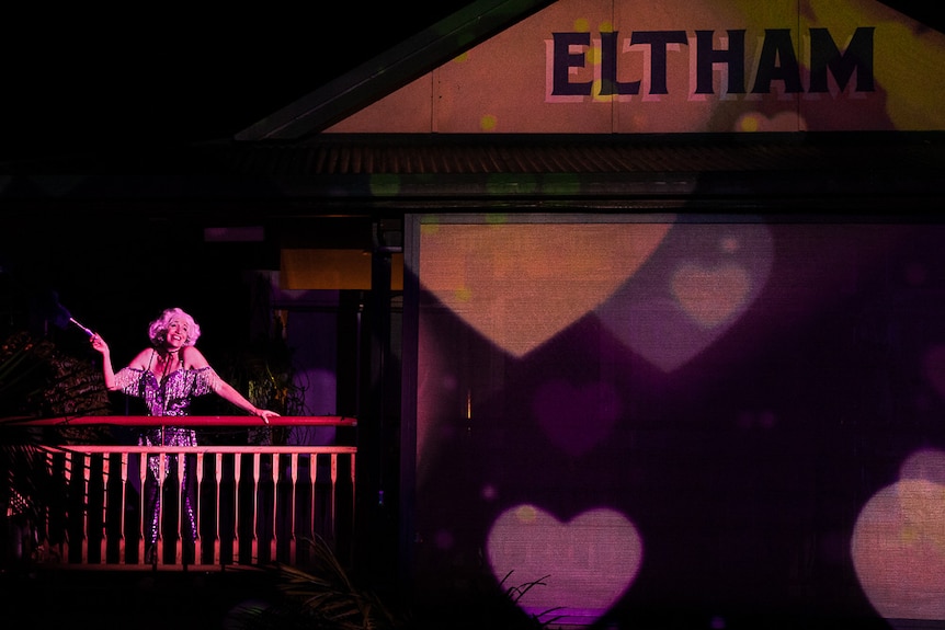 A woman in a beaded dress and blonde wig performs on the balcony of a historic pub with the word Eltham on the roof above her.