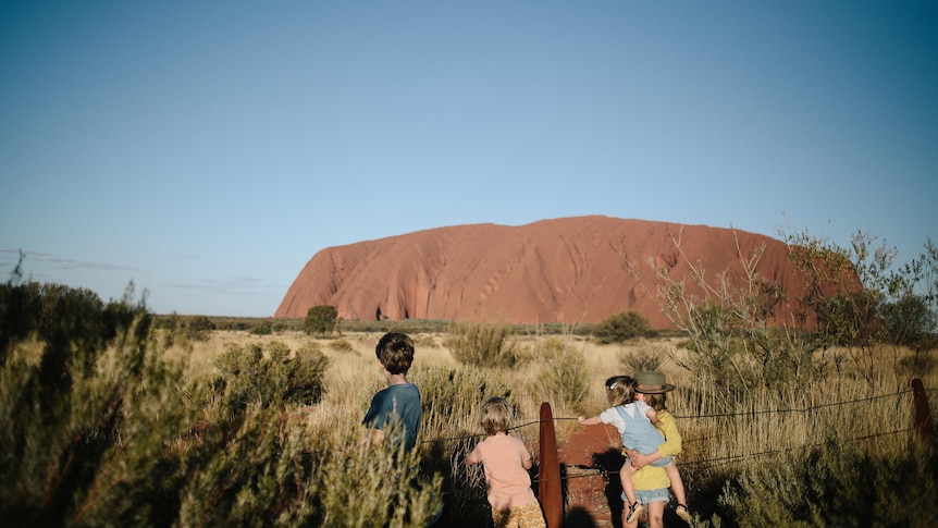 Four young children seen from behind at a wire fence with an expanse of blue sky and the huge red Uluru behind them.