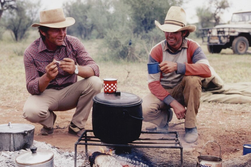 Two men holding cups crouch by a bit pot on a campfire.