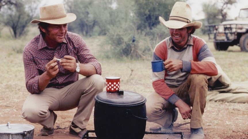 Two men holding cups crouch by a bit pot on a campfire.