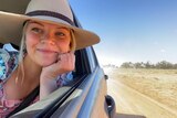 Woman wearing akubra hat leans out of car window smiling on outback road