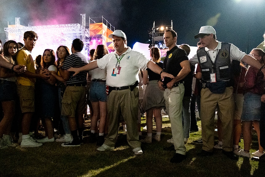 Two men wearing cargo pants and white polo and caps hold back crowd at outdoor night time event in a grass area.