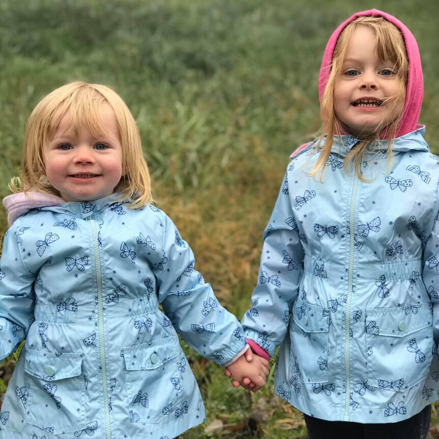 The two little girls have blonde hair and are wearing blue jackets, holding hands and smiling outside.