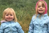 The two little girls have blonde hair and are wearing blue jackets, holding hands and smiling outside.