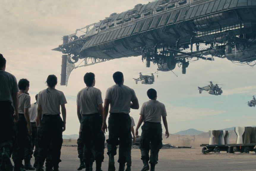 Solders are seen peering up at a spaceship on a cloudy day. They wear white shirts and cargo pants and are silhoutted.