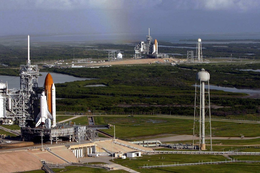 Two space shuttles on launchpads: one rocket in the foreground, the other in the distance, with trees and lakes between them.
