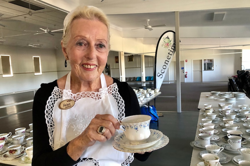 A lady wearing a maids uniform holds up a cup an saucer, with more cups and saucers on tables in the background.