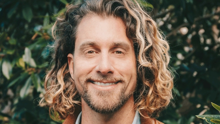 A person in a brown jacket against a tree smiles in a profile photograph.