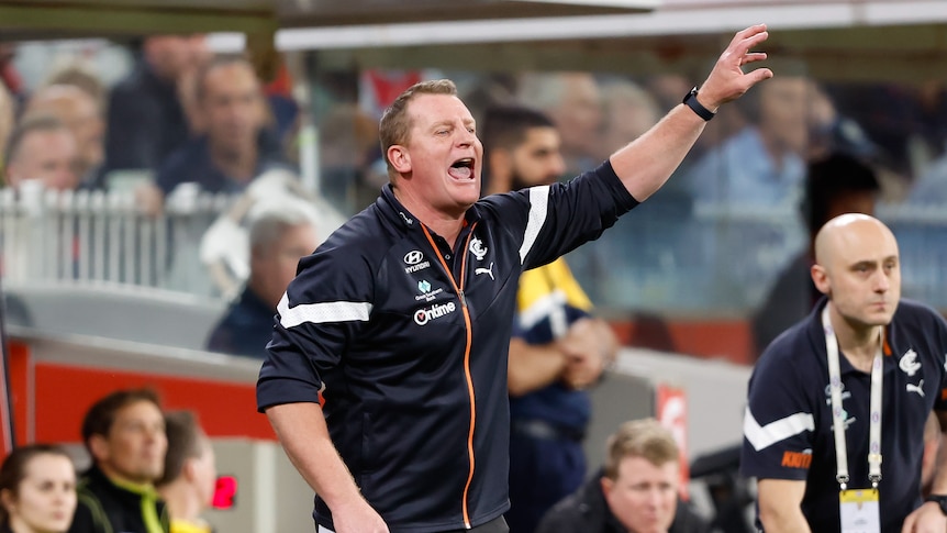 A Carlton AFL coach shouts to his players on the field during a final while holding one hand in the air.