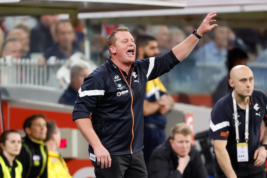 A Carlton AFL coach shouts to his players on the field during a final while holding one hand in the air.