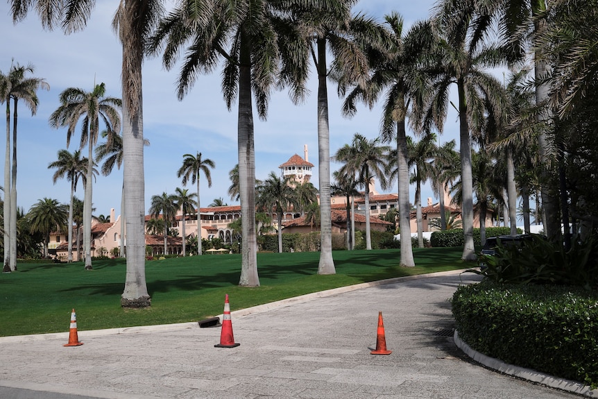 A white and pink-ish mansion lies behind a lush green lawn and driveway lined with palm trees.