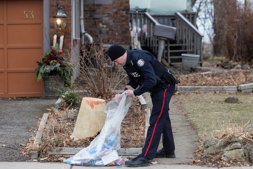 A police officer bends down and looks into a plastic bag of rubbish outside a house where bodies were found in planters.