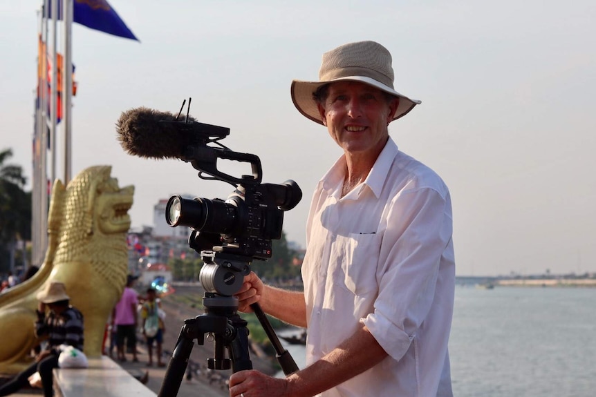 Cameraman and camera standing on edge of river with Cambodia street scene in background.
