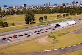 An aerial photo of cars lining up in two rows behind two white tents. In the background is a city skyline