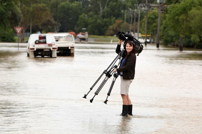 Woman wearing gumboots standing in floodwaters holding camera and tripod.