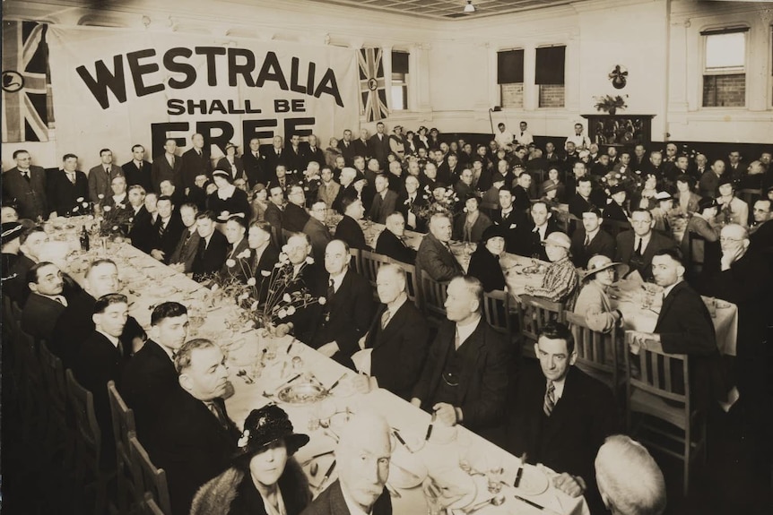 A sepia toned photograph shows dozens of men and women in 1934, sitting at several long tables in a large hall