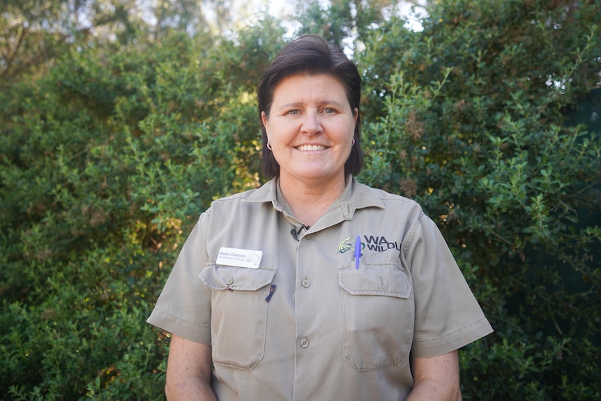 A woman wearing a beige shirt stands in front of a green bush