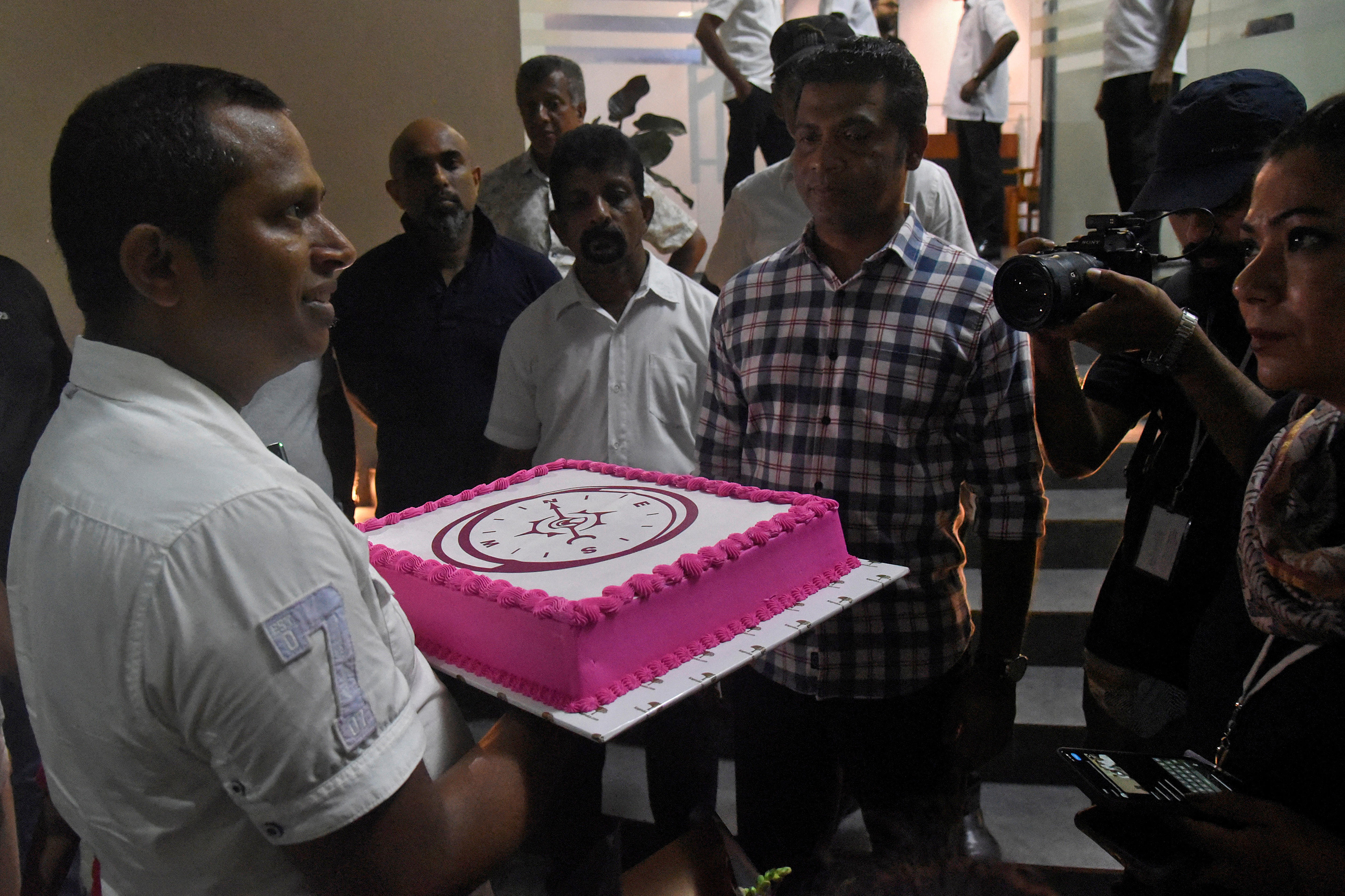 A group of men stand in a circle in front of a camera, with one man holding a white and pink cake