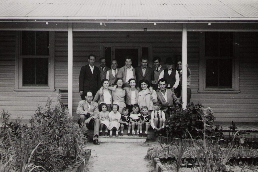 Old black and white photograph of a group of men, women and children posing for a photo on the steps of a small wooden home.