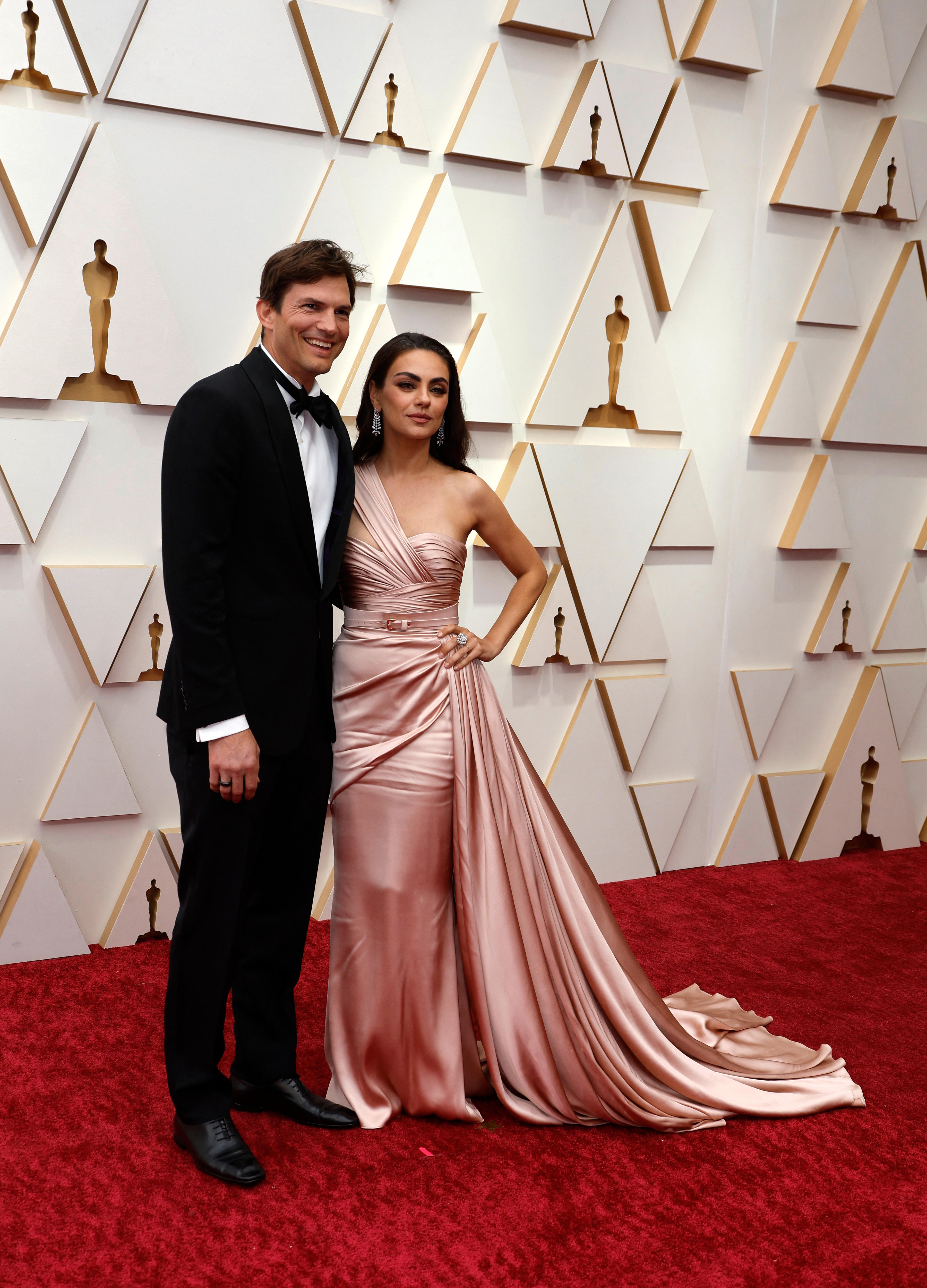 couple ashton kutcher and mila kunis stand arm in arm on the red carpet. kunis wears a dusty pink silk gown with a side train