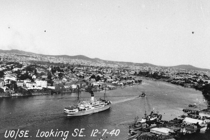 A black and white aerial photo above a river. A tug guides a ship along the Brisbane River. 