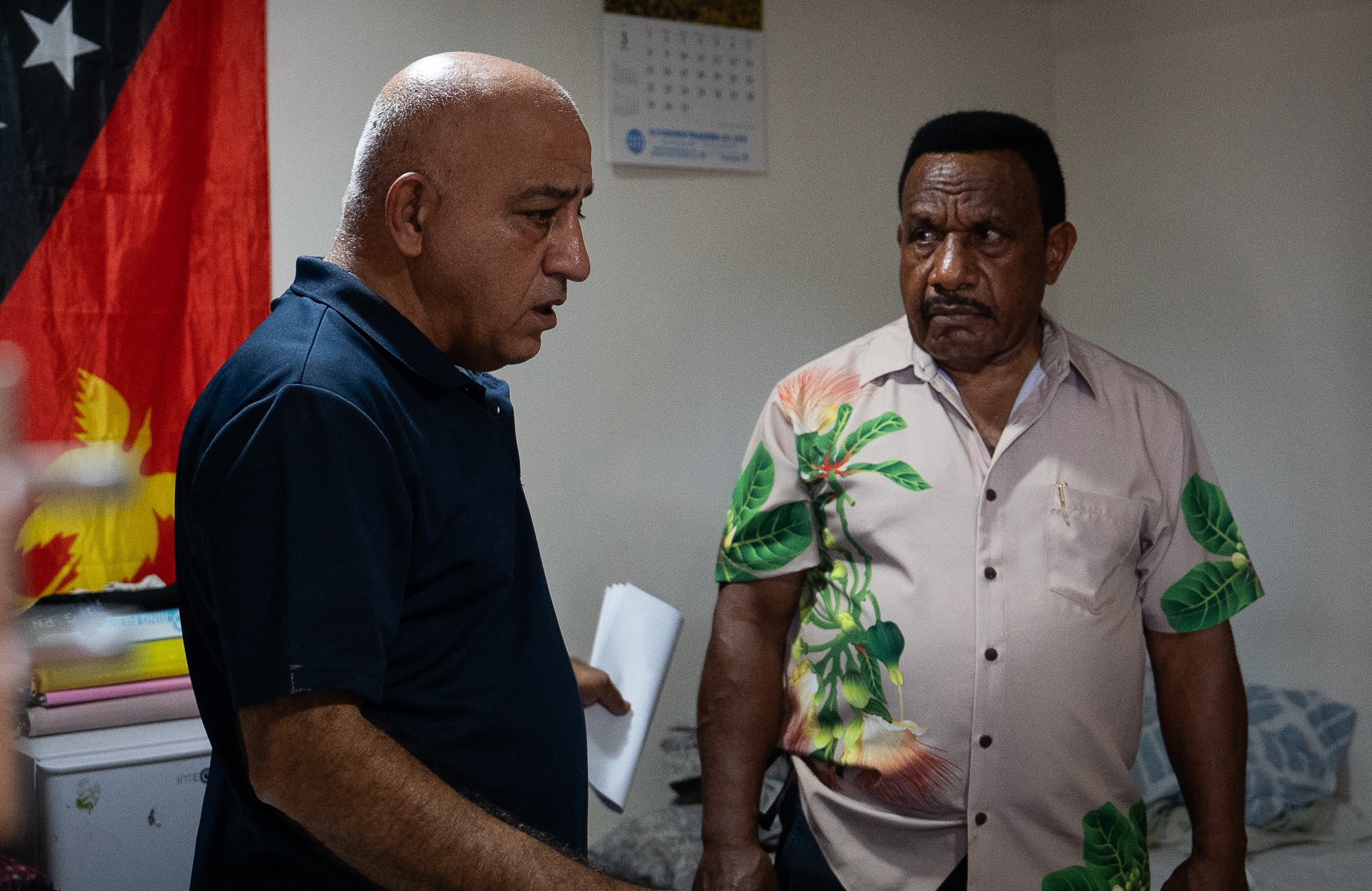 Two middle-aged men, one in a dark shirt and the other in a floral-patterned button-up, stand in a hotel room.