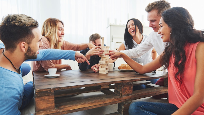 A diverse group of young people around a wooden table play a wooden stacking game while laughing together. 