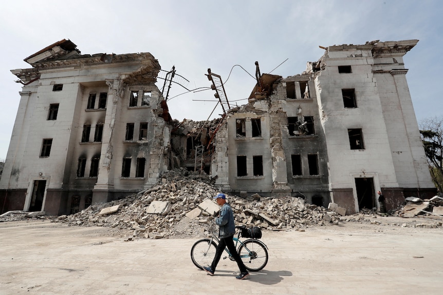 A person on a bike walks in front of a destroyed building with rubble.