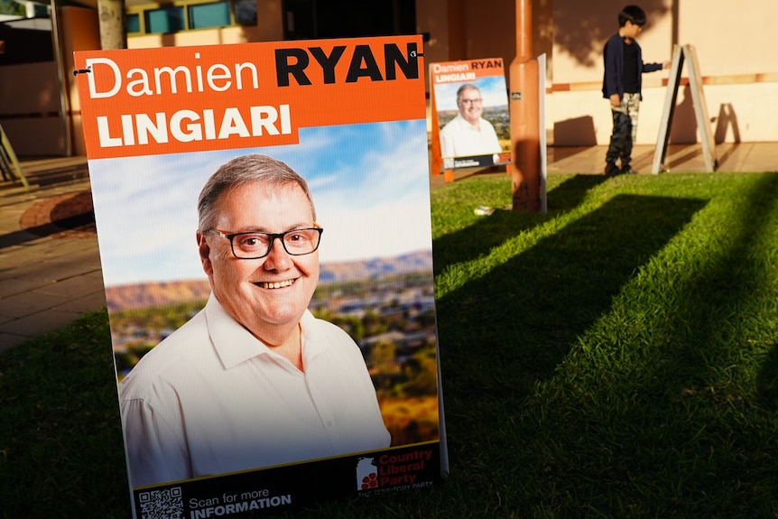A poster reading 'Damien Ryan Lingiari', with an image of a smiling manwith glasses on it, stuck in grass in front of a building