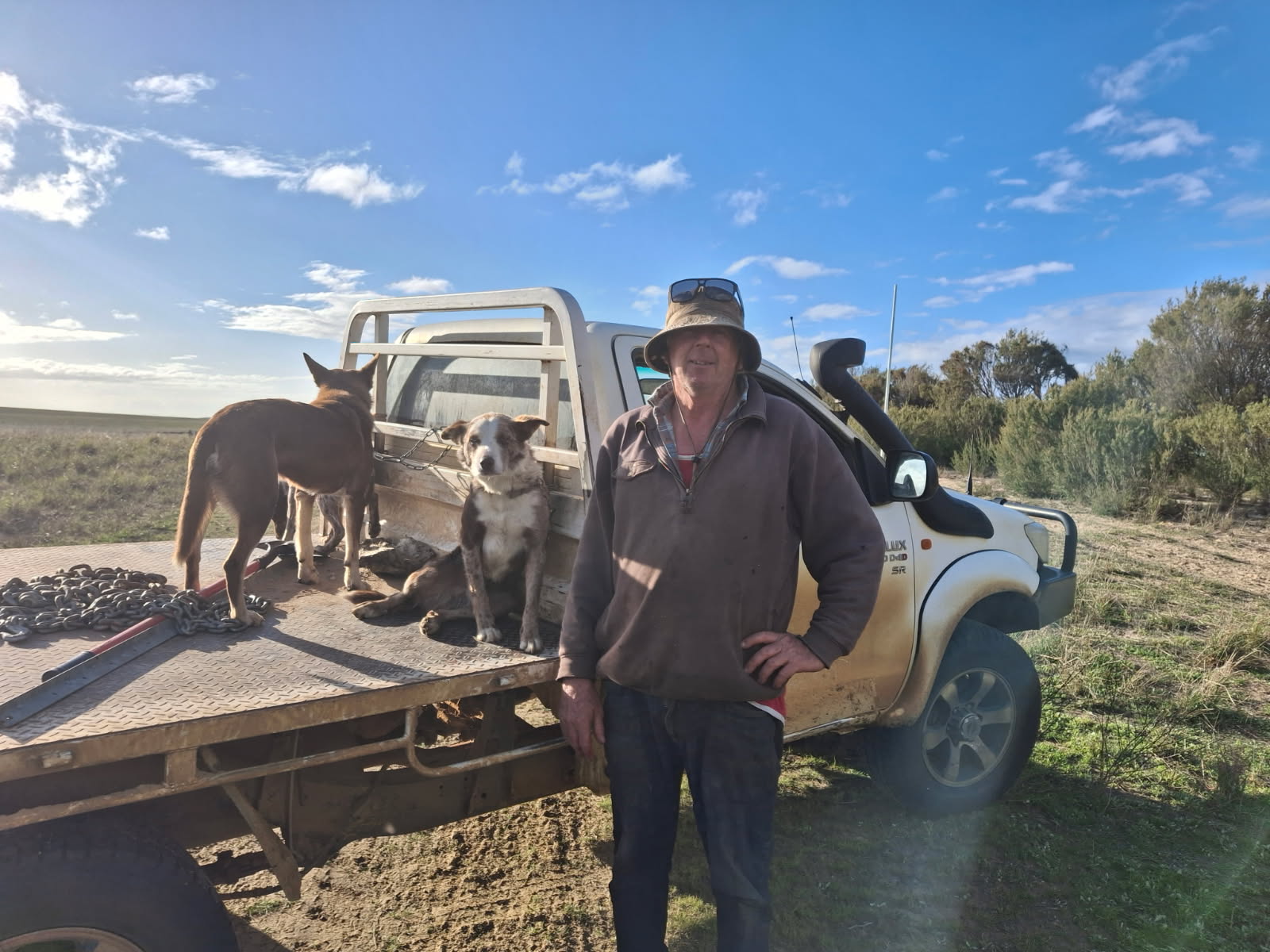 A man in a hat and jumper stands in front of his ute in a paddock. A couple of working dogs are on the tray.