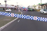 Behind police tape are two ambulances with lights flashing parked on a road with a few other vehicles nearby