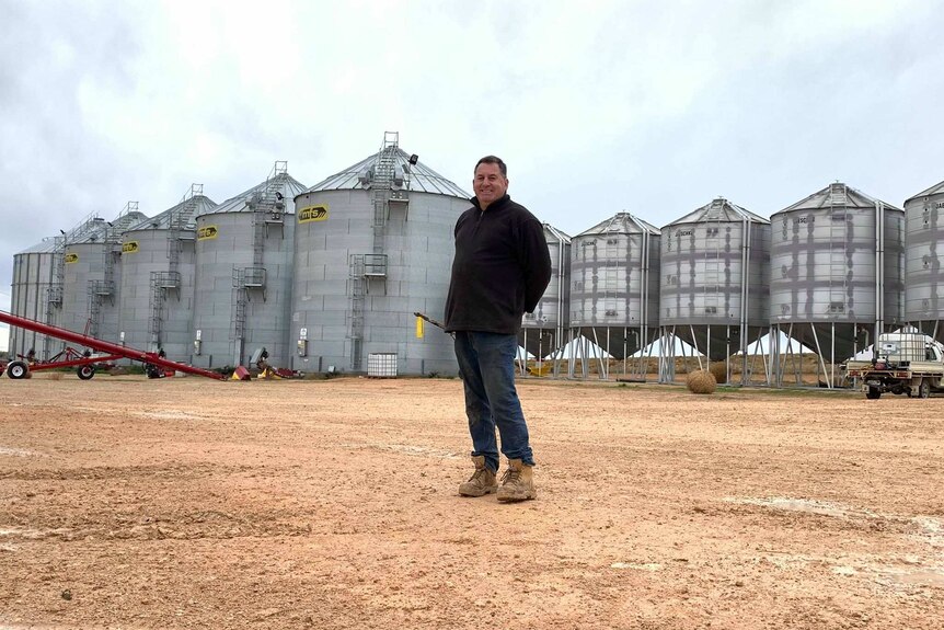 Corey Blacksell, a white man, stands in front of several large silver grain silos.