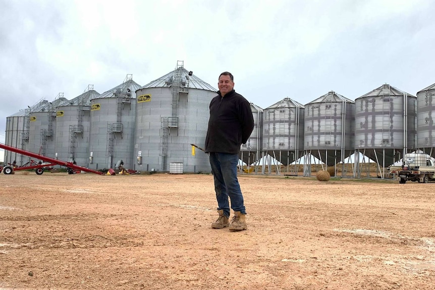A smiling man stands in front of grain silos in a rural town
