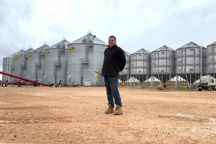 Corey Blacksell, a white man, stands in front of several large silver grain silos.