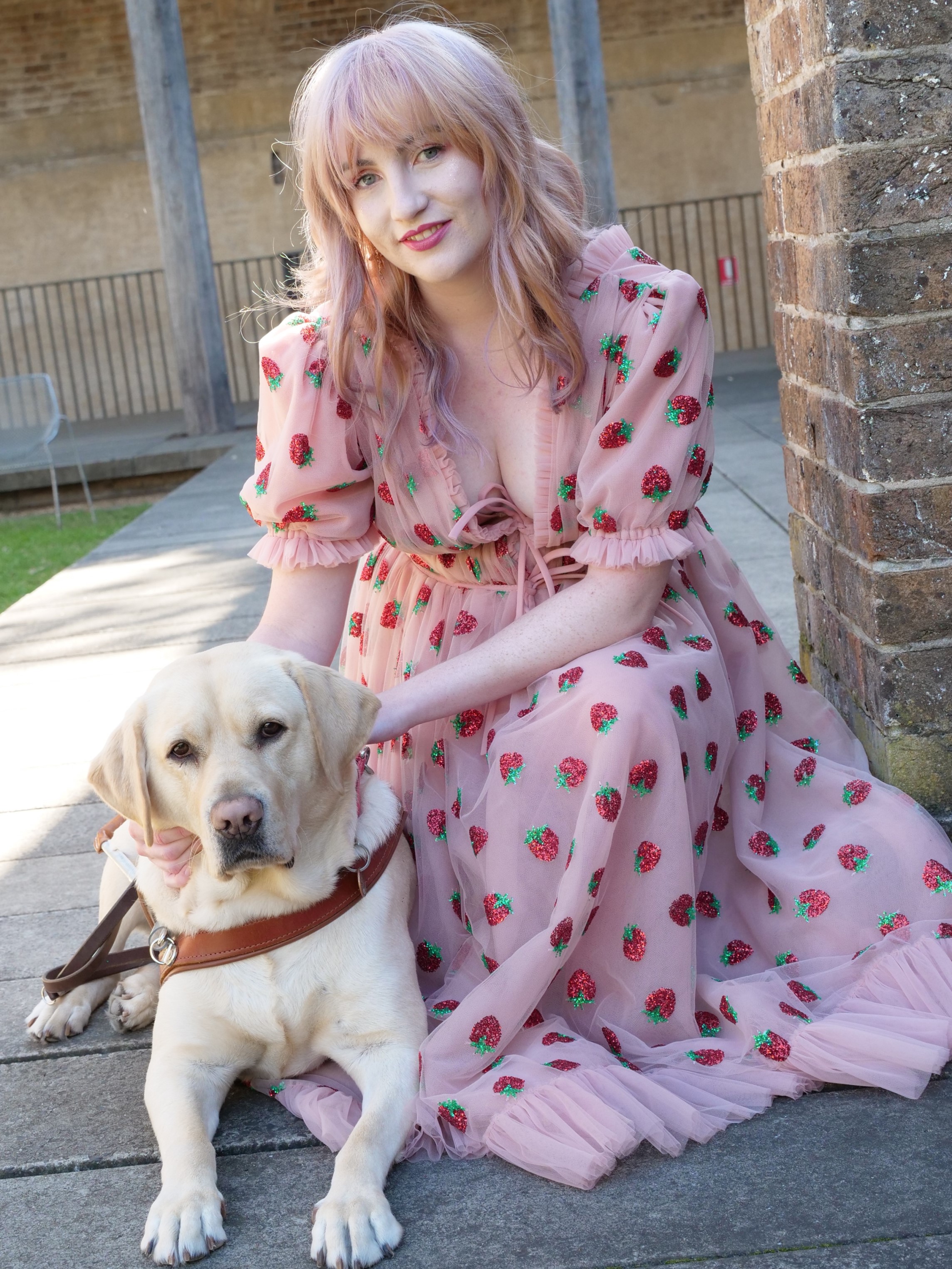 Ingrid kneels behind her Guide Dog, Banner, who is sitting,  wearing a pink dress with strawberries.