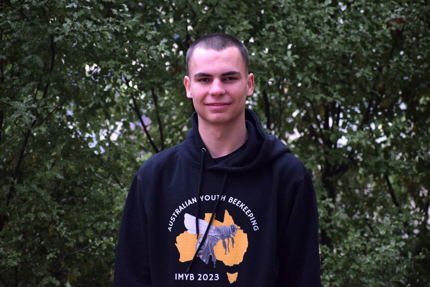Teen standing smiling at camera, shaved head, wearing black hooded jumper with Australia map and bee on front.