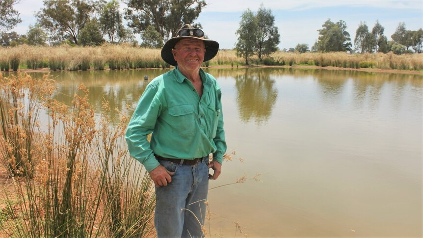 A farmer smiles to the camera in front of a dam, with native plants surrounding it.