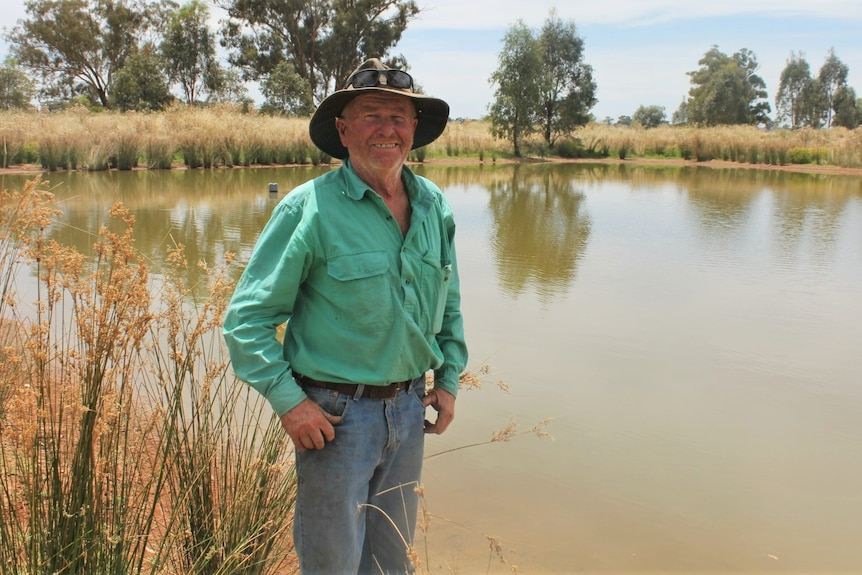 A farmer smiles to the camera in front of a dam, with native plants surrounding it.