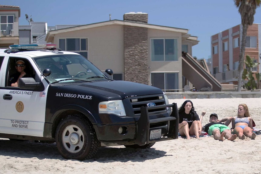 A San Diego Police vehicle sits on the beach in front of three people lying on the sand.