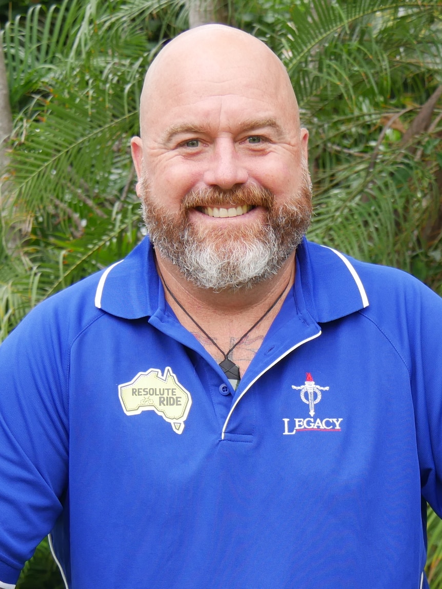 Man in blue shirt stands behind his bicycle in front of trees.