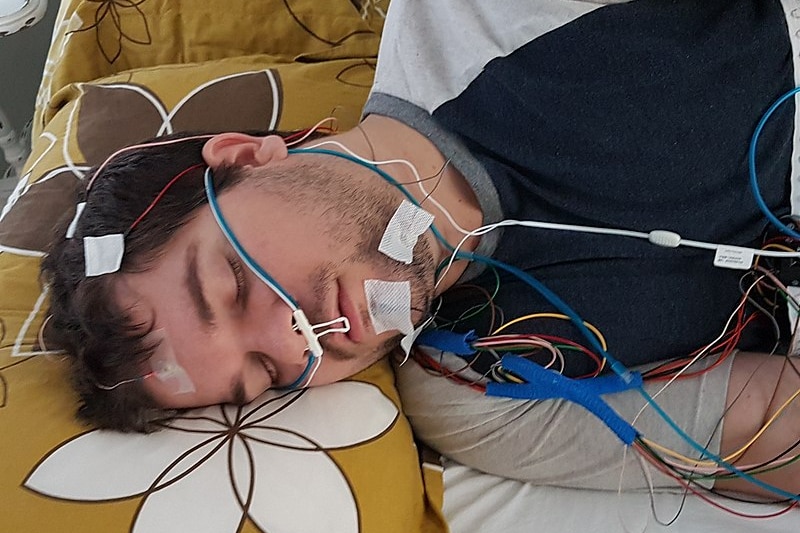 A man sleeps on his side with multicoloured wires and tubes taped to his head and face