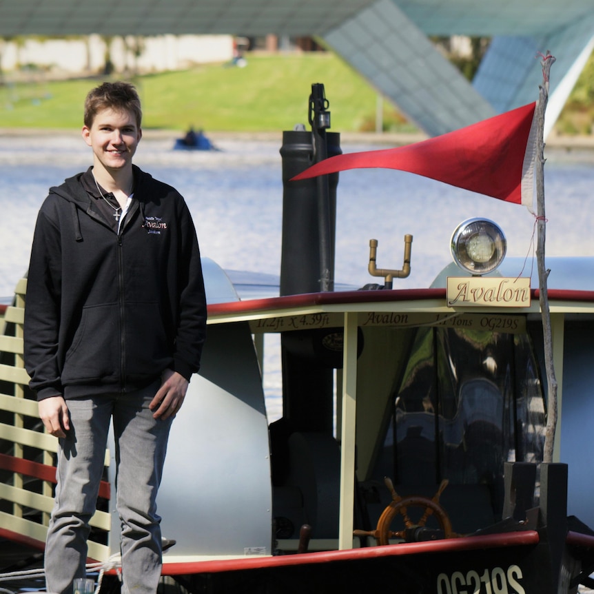 young man in blue top and jeans standing in front of a paddle steamer, with a river in the background.