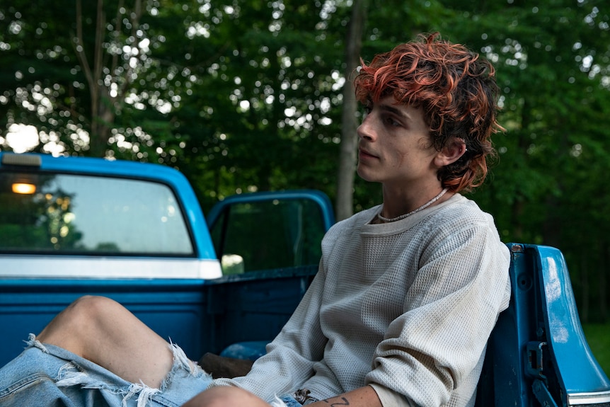 A pale young man with dyed red hair and ripped jeans sits in the trailer of a blue truck