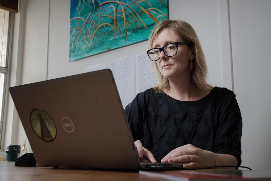 Woman with blonde hair wearing a black top and glasses looking at a laptop.
