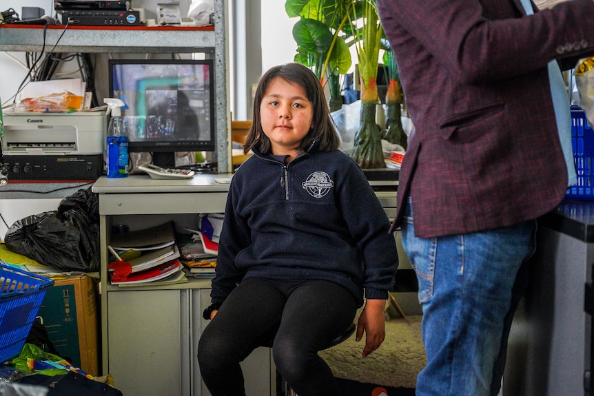 A young girl in a school uniform sits on a high stool behind a cash register.