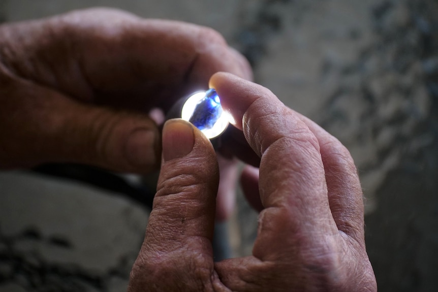 Aged hands hold a torch light under a precious blue gemstone.