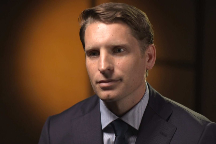 Head shot of Andrew Hastie wearing grey suit and dark tie, with circle of orange light on the wall behind him