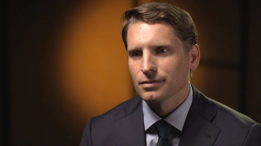 Head shot of Andrew Hastie wearing grey suit and dark tie, with circle of orange light on the wall behind him